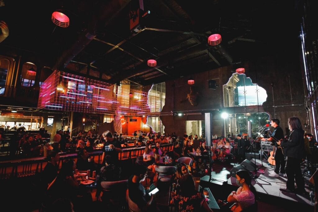 ภาพบรรยากาศสถานที่จัดปีใหม่ภายในร้านอาหารและบาร์ที่มีวงดนตรีสดแสดงบนเวที ผู้ร่วมงานนั่งสังสรรค์อย่างเป็นกันเอง ท่ามกลางแสงไฟยามค่ำคืน เหมาะสำหรับงานเลี้ยงส่งท้ายปี งานบริษัท และการฉลองปีใหม่ที่ต้องการทั้งความบันเทิงและประสบการณ์พิเศษ