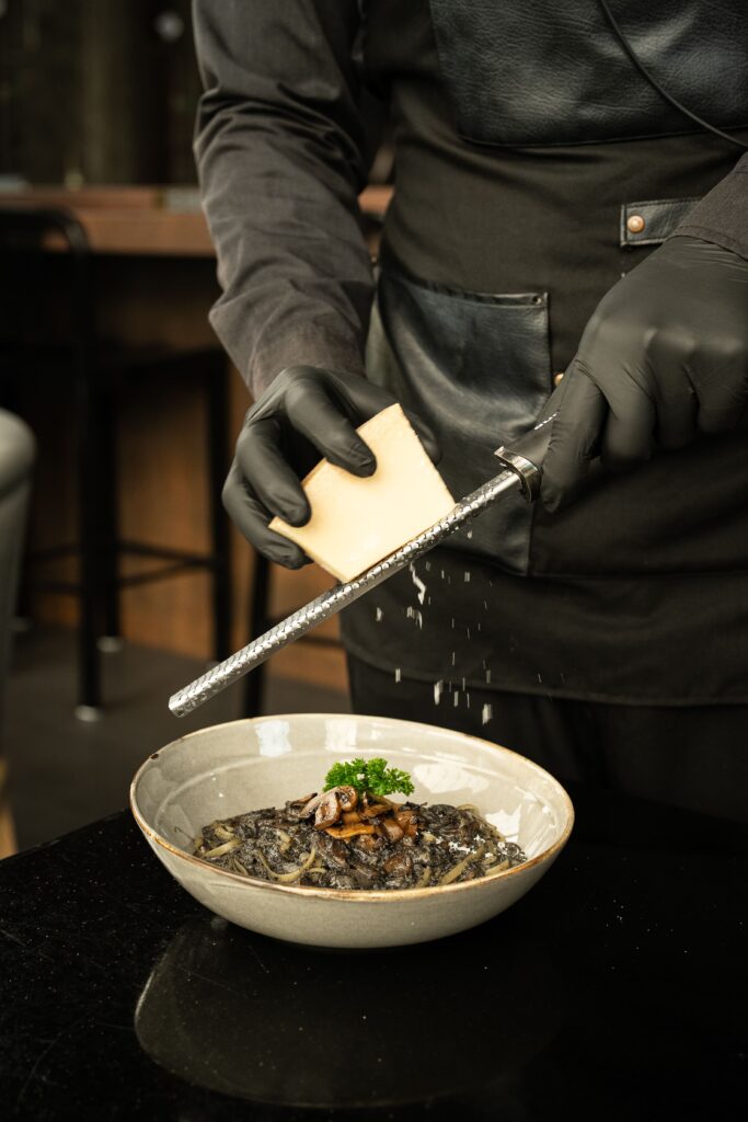 เชฟกำลังขูดชีสสดบนพาสต้าเส้นดำซิกเนเจอร์ที่ DAVIN CAFE ร้านอาหาร เลียบด่วน โดดเด่นด้วยการเสิร์ฟอาหารระดับพรีเมียมและประสบการณ์ Fine Dining ที่แตกต่าง