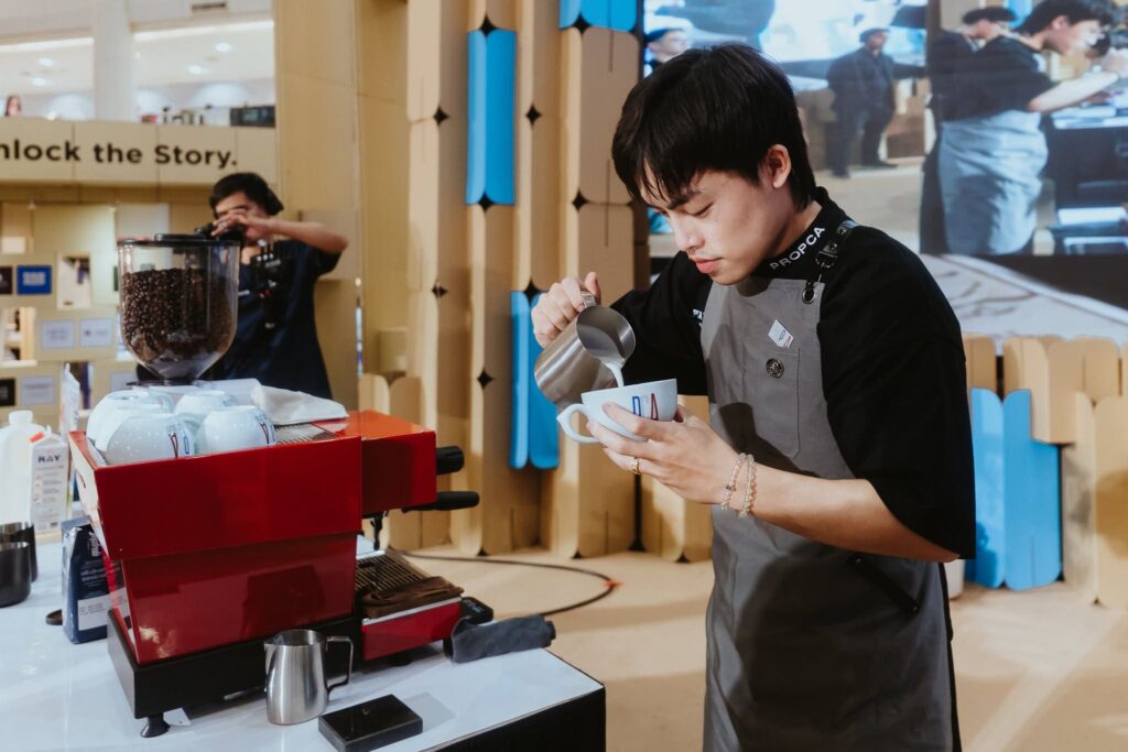 บาริสต้า DAVIN CAFE กำลังแข่งขันสร้างสรรค์ลาเต้อาร์ตบนเวที PCA Latte Art Thailand 2026 แสดงความแม่นยำในการเทนม ทักษะด้านกาแฟพิเศษ และมาตรฐานการแข่งขันบาริสต้าระดับประเทศท่ามกลางอุปกรณ์กาแฟมืออาชีพ