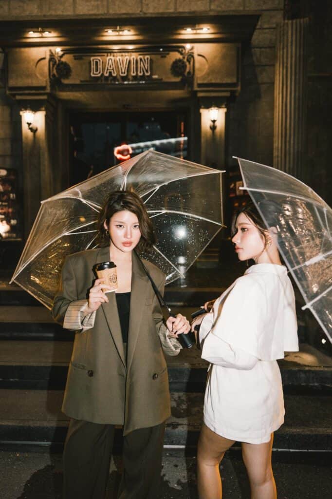 Cafe photo poses with two women holding clear umbrellas and coffee cups at the DAVIN CAFE entrance in a rainy fashion editorial style